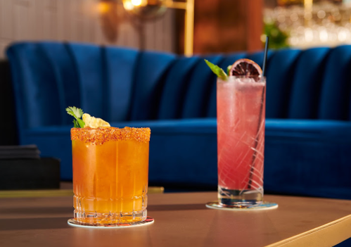 Two colorful cocktails on a table: one orange with a rim of spice, the other pink with a garnish, set against a blue backdrop.
