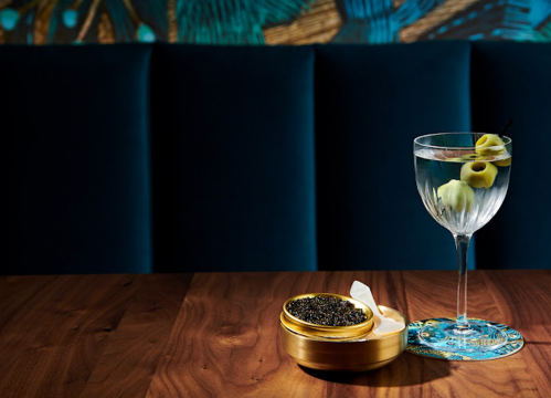 A glass of martini with olives sits beside a tin of caviar on a wooden table, against a plush blue backdrop.