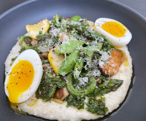 A bowl of creamy polenta topped with sautéed greens, mushrooms, lemon slices, and soft-boiled eggs, garnished with cheese.