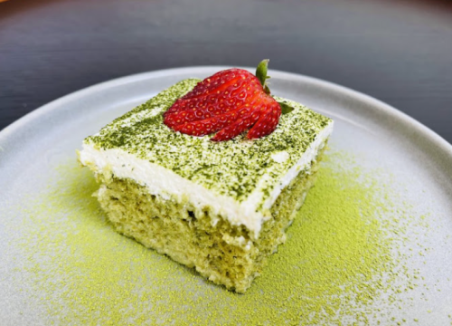 A slice of matcha cake topped with whipped cream and a fresh strawberry, served on a light-colored plate.