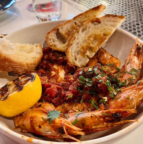 A bowl of shrimp in tomato sauce, garnished with herbs, served with grilled lemon and slices of toasted bread.