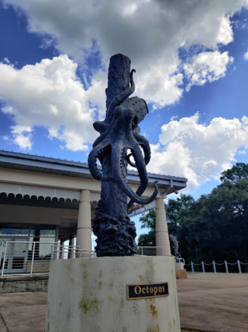 A bronze octopus sculpture wraps around a tall pedestal under a blue sky with fluffy clouds.