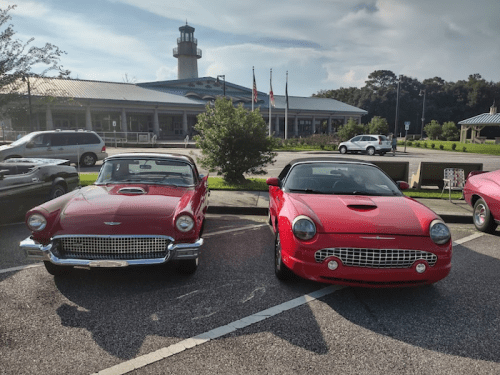 Two classic red cars parked side by side in front of a building with a lighthouse and flags in the background.
