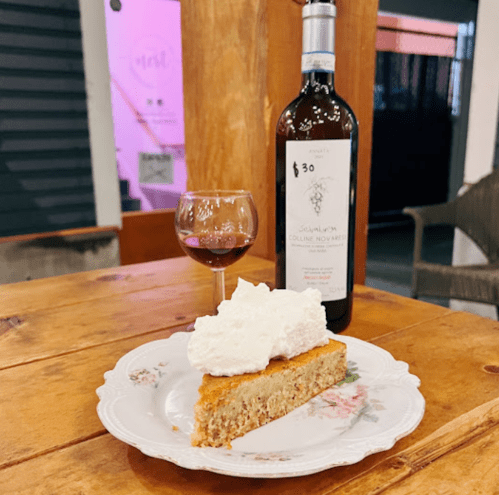 A slice of cake topped with whipped cream, a glass of red wine, and a bottle of wine on a wooden table.