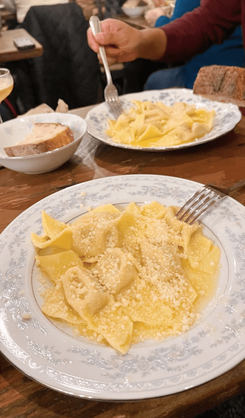Two plates of pasta topped with cheese, accompanied by a slice of bread and a drink in the background.