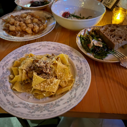 A table setting with plates of pasta, gnocchi, greens, and bread, illuminated by a soft candlelight.