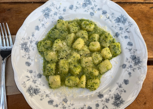 A plate of gnocchi in a green pesto sauce, garnished with grated cheese, on a floral-patterned dish. Forks beside it.