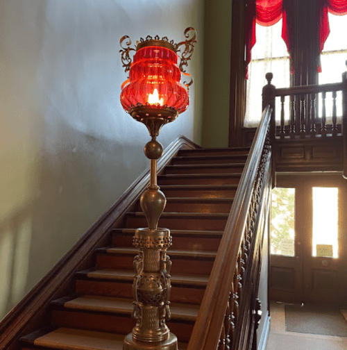 A vintage lamp stands beside a wooden staircase, with red curtains and sunlight streaming through a nearby door.