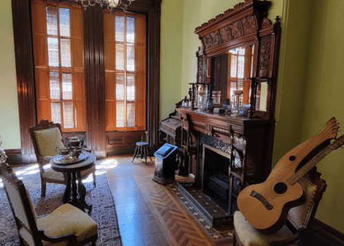 A vintage room with wooden furniture, a fireplace, a guitar, and large windows letting in natural light.