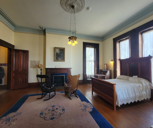 A spacious vintage bedroom with a wooden bed, rocking chairs, a fireplace, and large windows letting in natural light.