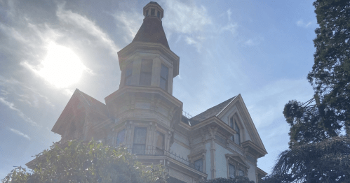 Historic Flavel House Museum with a tall turret, surrounded by greenery, under a bright sky. Built in 1885.