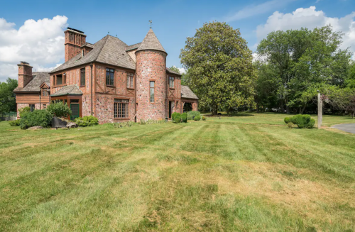A large brick house with a turret, surrounded by a manicured lawn and trees under a blue sky with clouds.