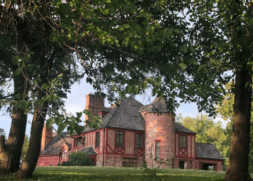 A historic brick house with a unique design, surrounded by trees and greenery under a clear sky.