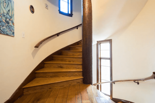 Curved wooden staircase with a smooth railing, leading to a bright windowed landing. Wall art visible on the left.