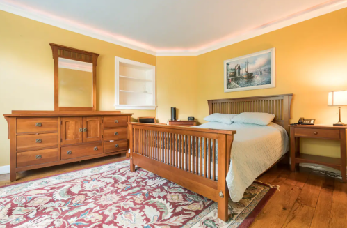 Cozy bedroom with a wooden bed, dresser, and nightstand, featuring warm yellow walls and a decorative rug.