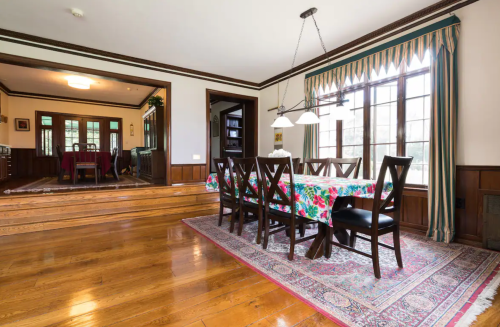 A spacious dining room with a wooden table, floral tablecloth, and large windows, featuring hardwood floors and elegant decor.