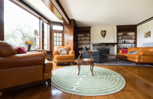 Cozy living room with leather sofas, a round rug, a coffee table, and a fireplace, featuring large windows and bookshelves.