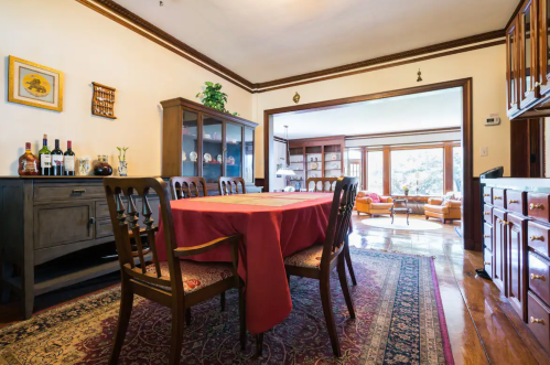 A dining room with a wooden table, chairs, and a cabinet, featuring a red tablecloth and a view into a living area.