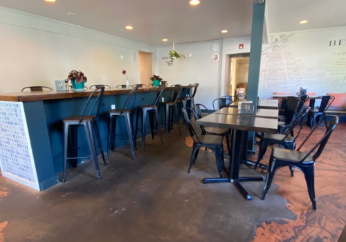 A cozy café interior featuring a bar with high stools and black tables surrounded by chairs. Warm lighting enhances the ambiance.