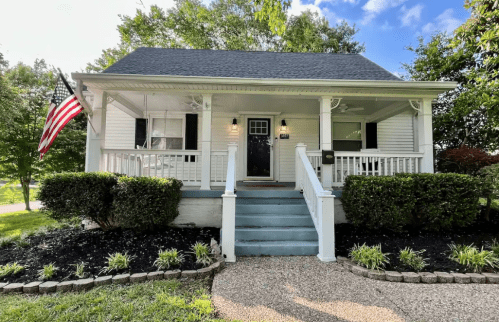 A charming white house with a front porch, steps, and an American flag, surrounded by greenery and landscaping.