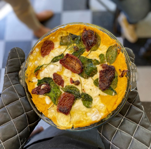 A baked dish with a golden crust, topped with spinach and sun-dried tomatoes, held in an oven mitt.