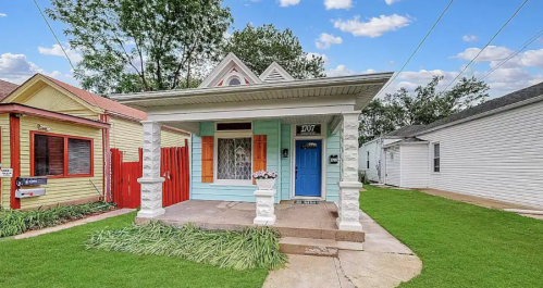 A charming blue house with a porch, surrounded by green grass and colorful neighboring homes under a blue sky.