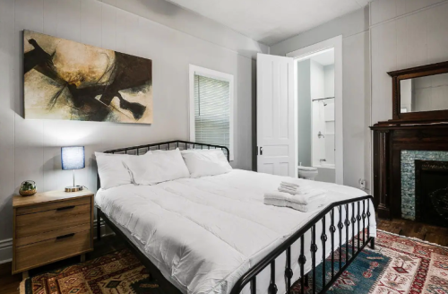 Cozy bedroom with a metal bed frame, artwork, nightstand, and an adjoining bathroom. Warm, inviting decor.