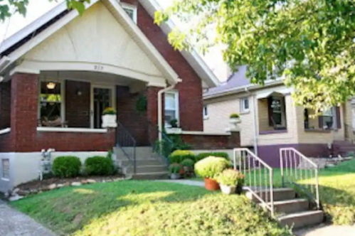 A charming brick house with a porch, surrounded by greenery and flower pots, next to a similar styled home.