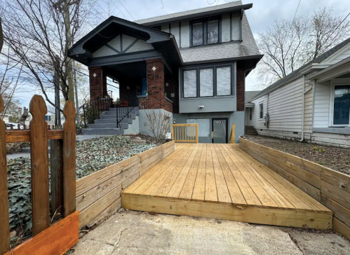 A wooden ramp leads to a two-story house with a porch, surrounded by trees and a wooden fence.