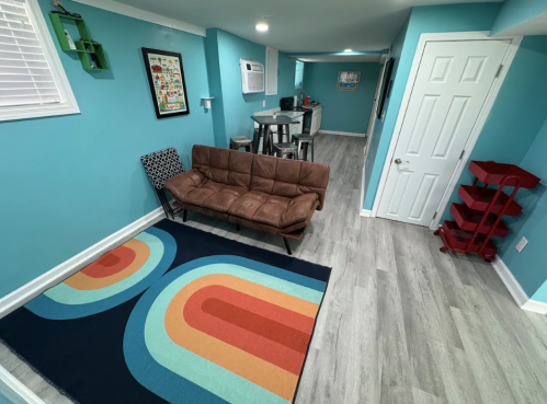 Cozy basement room with teal walls, a brown couch, colorful rug, and a small kitchenette area.