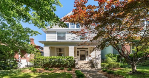 A charming two-story house with a front porch, surrounded by lush greenery and colorful trees on a sunny day.