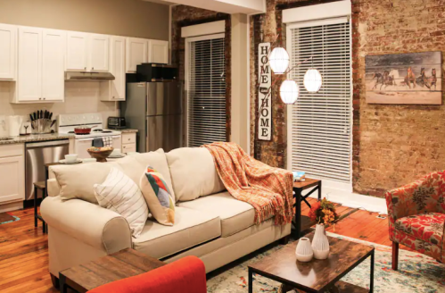 Cozy living room with a white sofa, colorful chairs, and a rustic brick wall, featuring warm lighting and decorative accents.