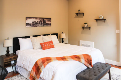 Cozy bedroom with a large bed, orange throw blanket, decorative pillows, and wall shelves.
