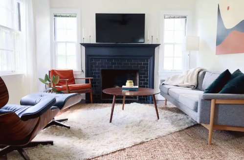 Cozy living room with a gray sofa, orange chair, coffee table, and a TV above a black fireplace, all on a plush rug.