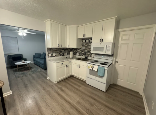 Modern kitchen with white cabinets, gray backsplash, and appliances; living room visible in the background.