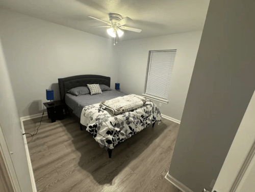 A cozy bedroom with a bed, nightstands, and a window, featuring light gray walls and wooden flooring.