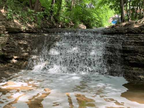 A small waterfall cascades over rocky ledges, surrounded by lush green trees and a serene natural setting.