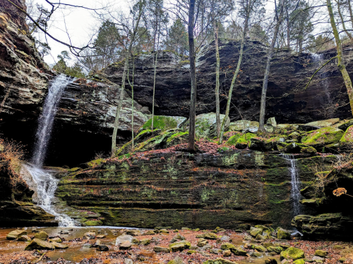 A serene landscape featuring two waterfalls cascading over rocky cliffs, surrounded by trees and autumn foliage.