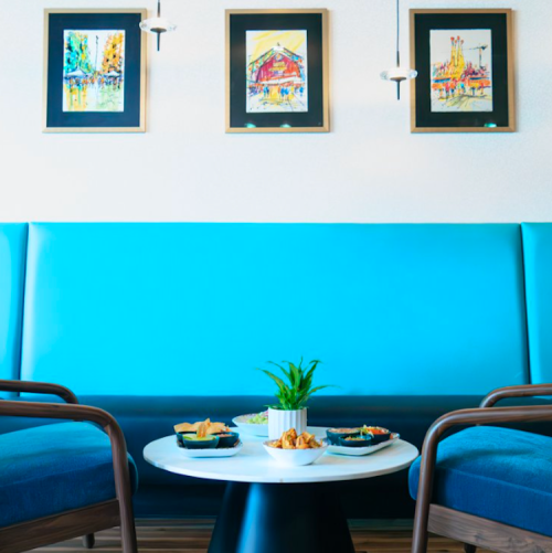 A bright seating area with blue benches, a round table, and colorful artwork on the walls. Snacks are on the table.