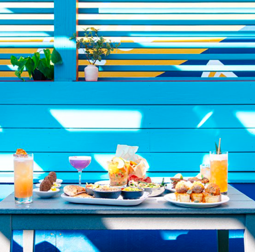 A vibrant table setting with colorful drinks and a variety of dishes against a blue and yellow backdrop.