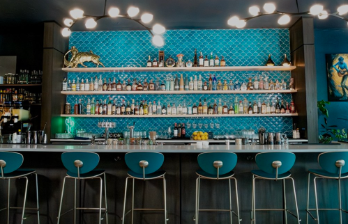 A stylish bar with a turquoise backsplash, shelves of liquor, and modern stools. Bright lighting adds a vibrant touch.