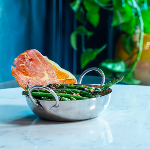 A silver bowl with green beans and a slice of crispy ham, set against a blue background with greenery.