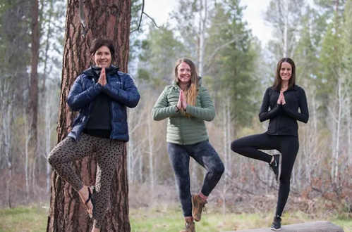 Three women practice yoga poses outdoors, surrounded by trees, with a serene natural backdrop.