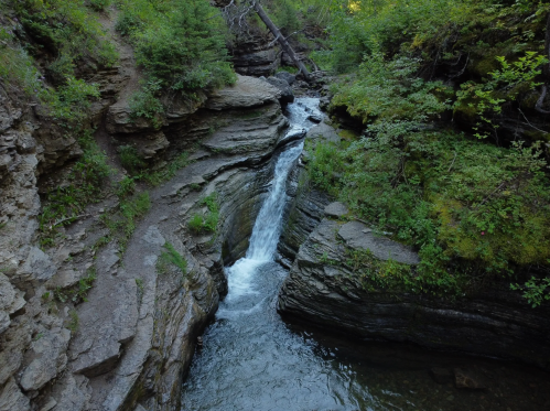 A serene waterfall cascades through rocky cliffs, surrounded by lush greenery and a tranquil stream below.