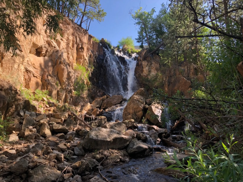 A scenic waterfall cascading down rocky cliffs, surrounded by trees and greenery under a clear blue sky.