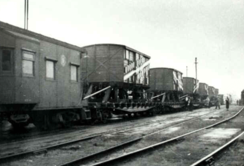 Black and white photo of train cars on tracks, with cargo containers and a figure standing nearby.