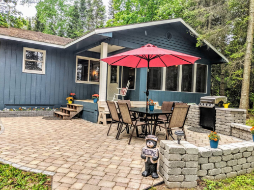 A cozy outdoor patio with a dining set, red umbrella, and grill, surrounded by greenery and flower pots.