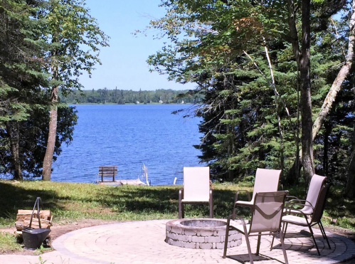 A serene lakeside view with a fire pit and chairs surrounded by trees on a sunny day.