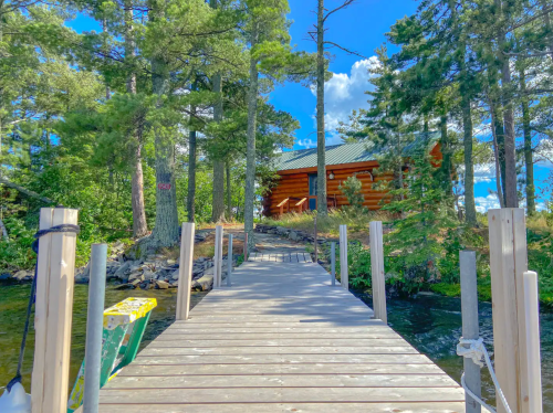 A wooden dock leads to a log cabin surrounded by tall trees and a clear blue sky.