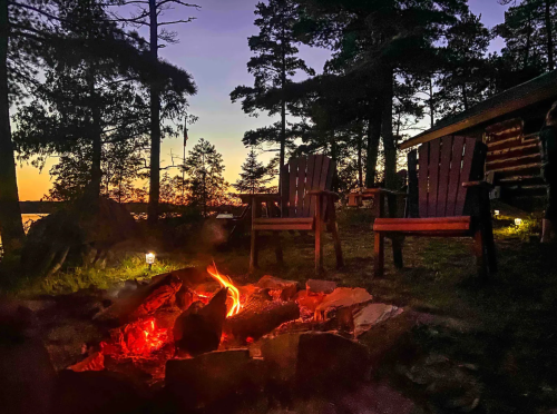 A cozy campfire scene at dusk, with two chairs facing the flames and a serene lake in the background.
